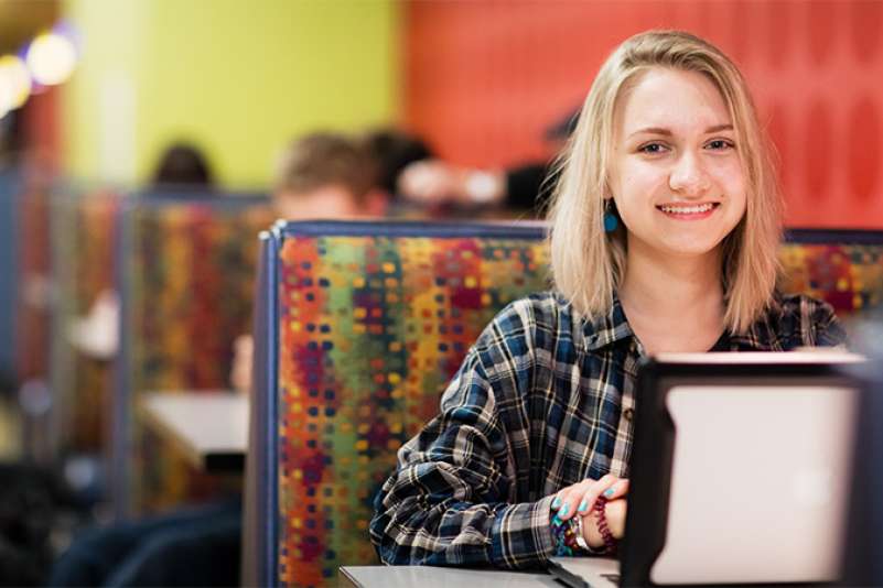 A student is sitting in the NTC cafeteria on her lapton with a confident smile