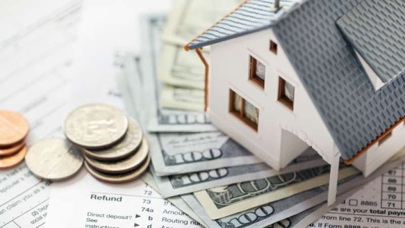 A small house model is set on top of several one-hundred dollar bills next to some coins.