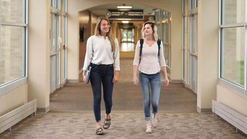 Two students walking down a hallway of the Wausau Campus, one carrying books, the other with a backpack on.