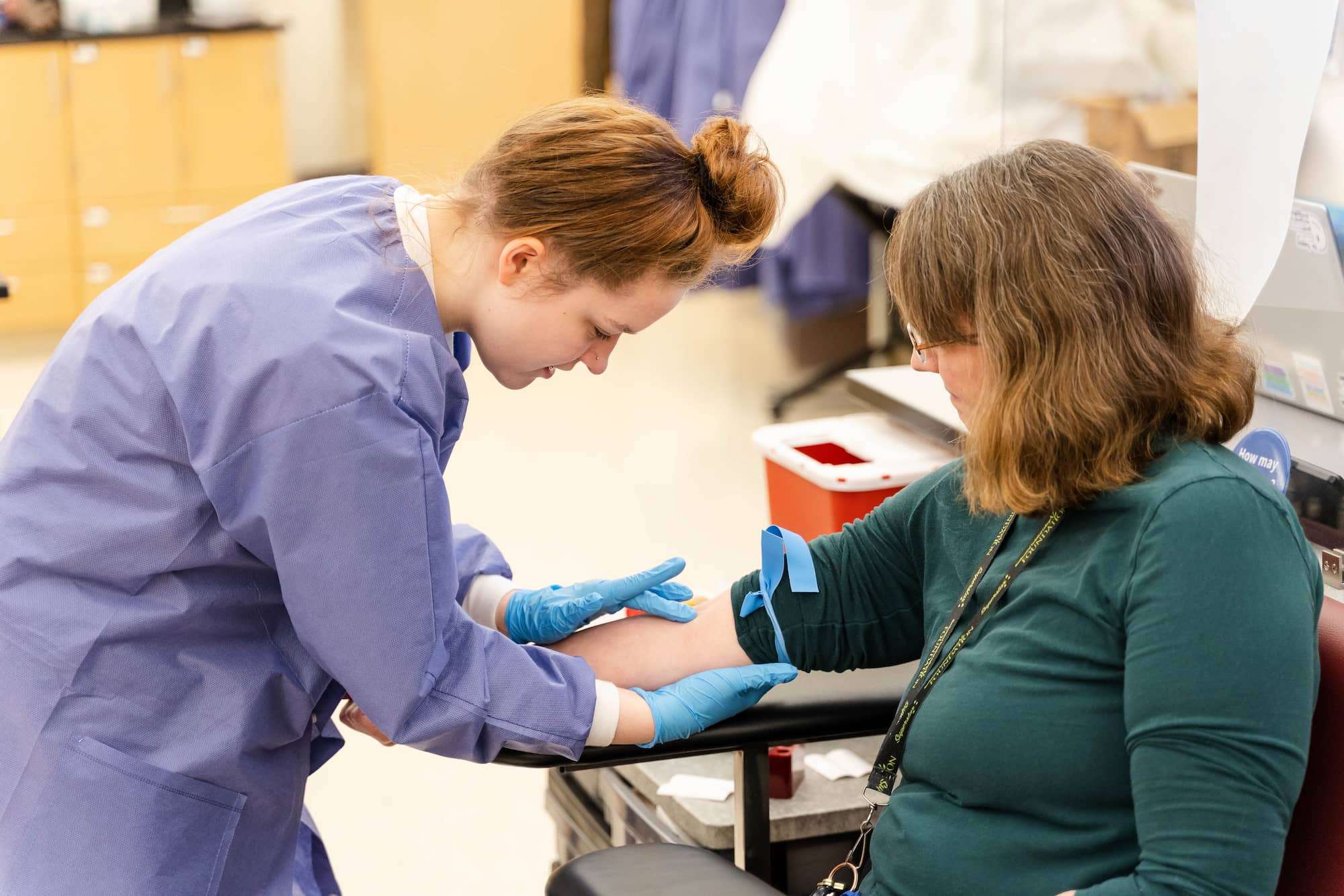 A student wearing rubber gloves presses gently with her fingers on a patient's forearm while finding the position of a vein.
