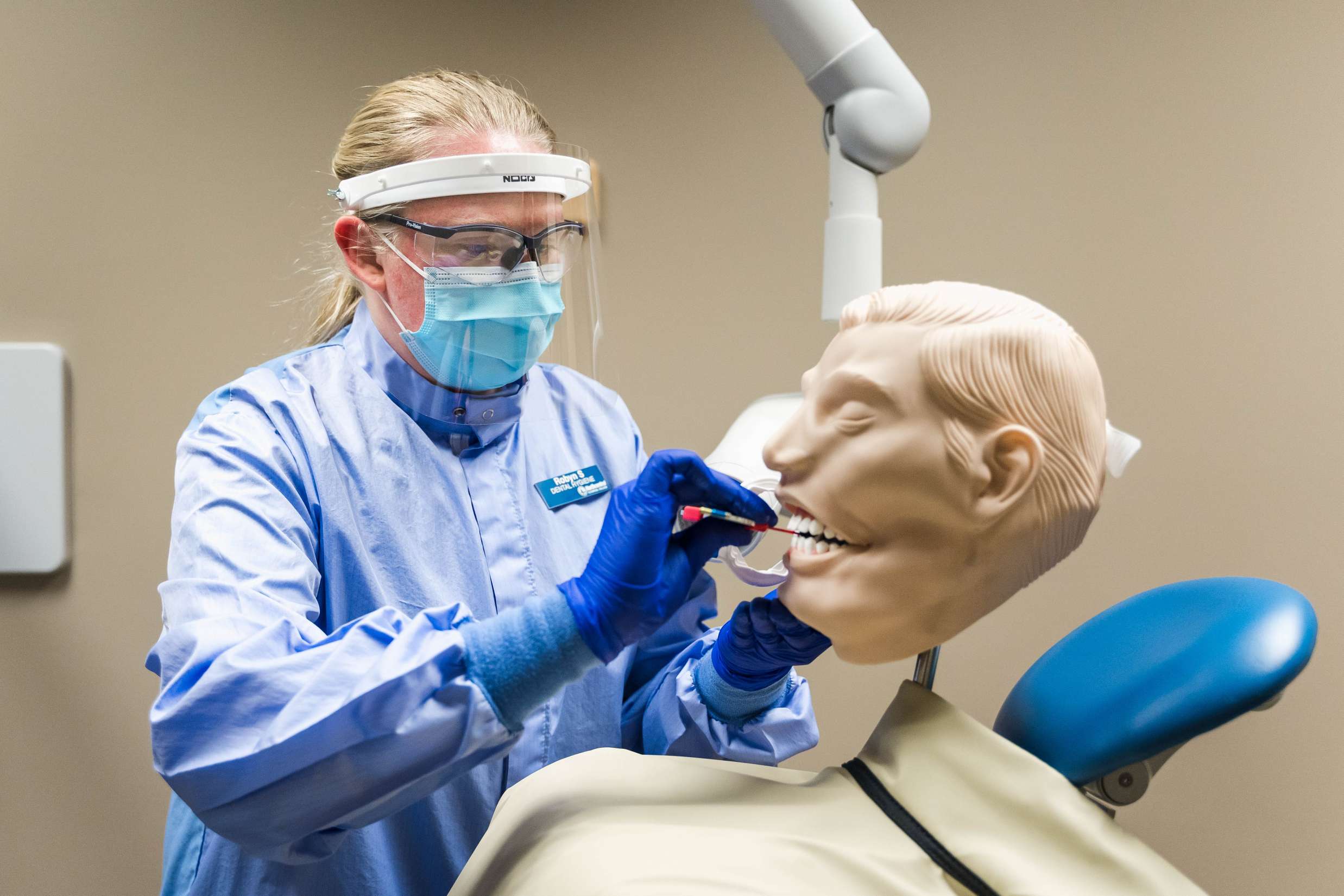 A dental hygienist performs oral cleaning procedures on a mannequin head positioned upright in a procedure room chair.