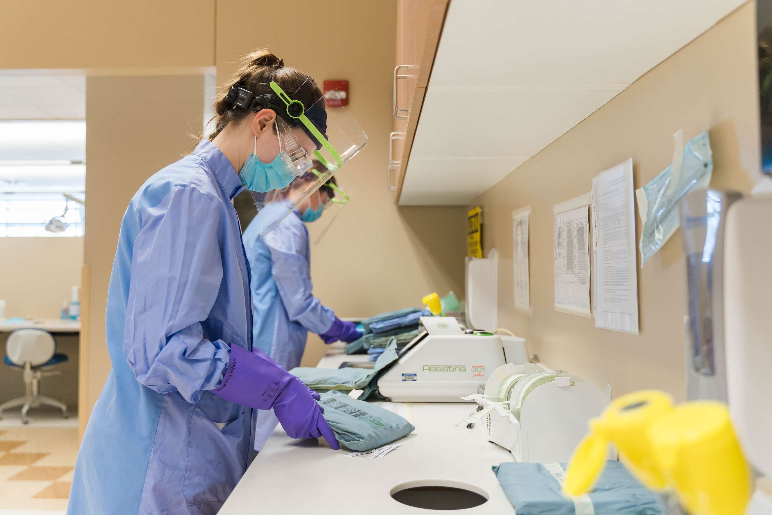 Two dental hygienists dressed in medical garbs wearing face shields working inside of a dental lab.