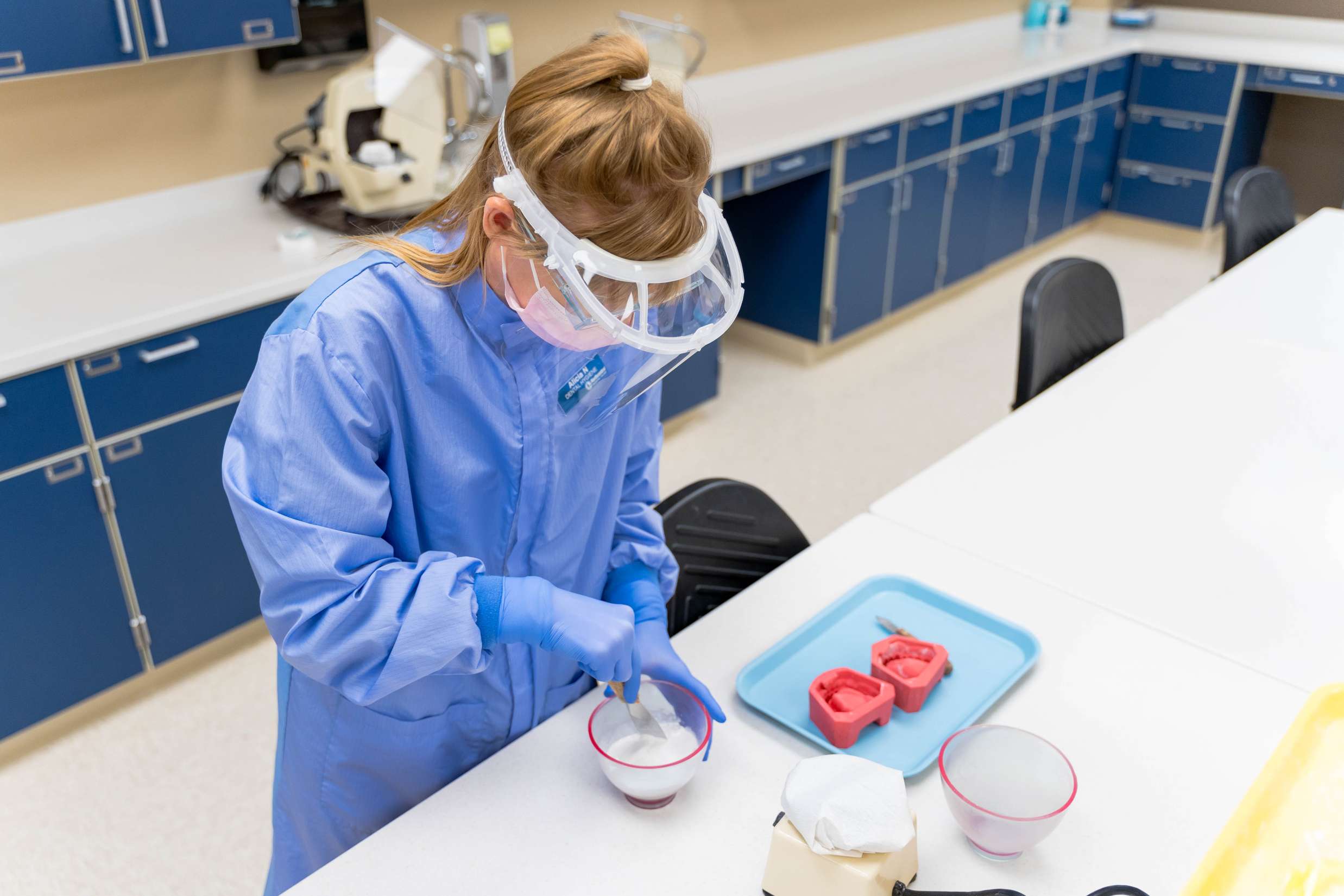 A dental Hygienist wearing blue medical garbs and a face shield is working with oral molds in a dental lab.