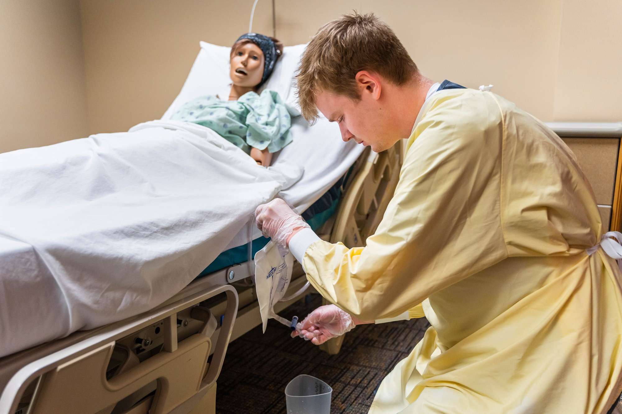 A Nursing Assistant empties out the contents of a colostomy bag into a container set on the floor next to a patient's bed.