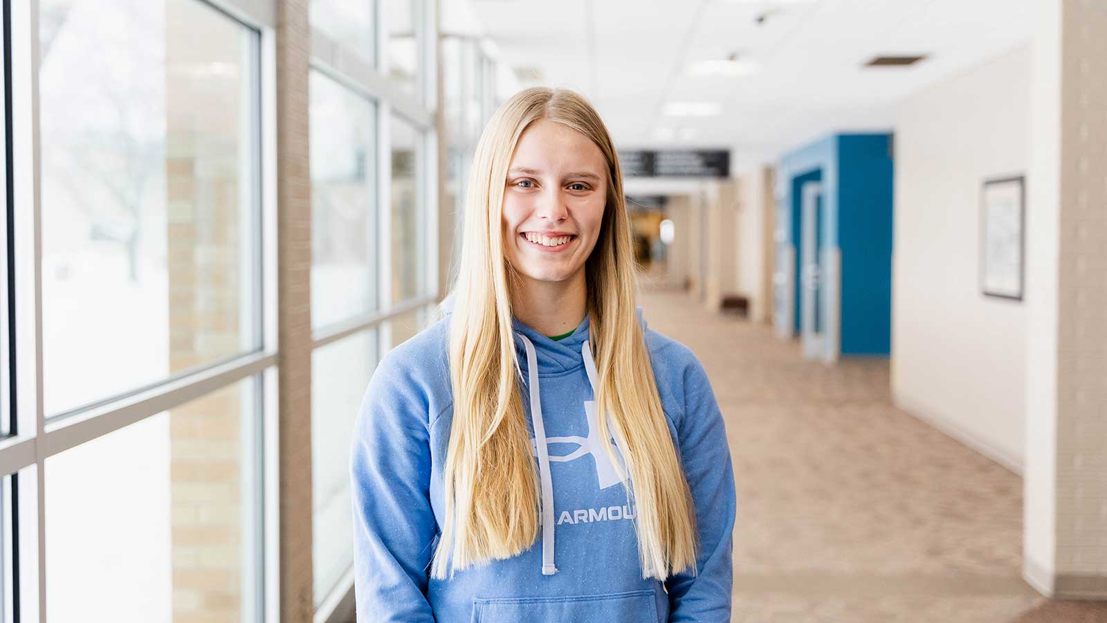 Ella standing in the hallway at NTC, smiling, while wearing a light blue hoodie.