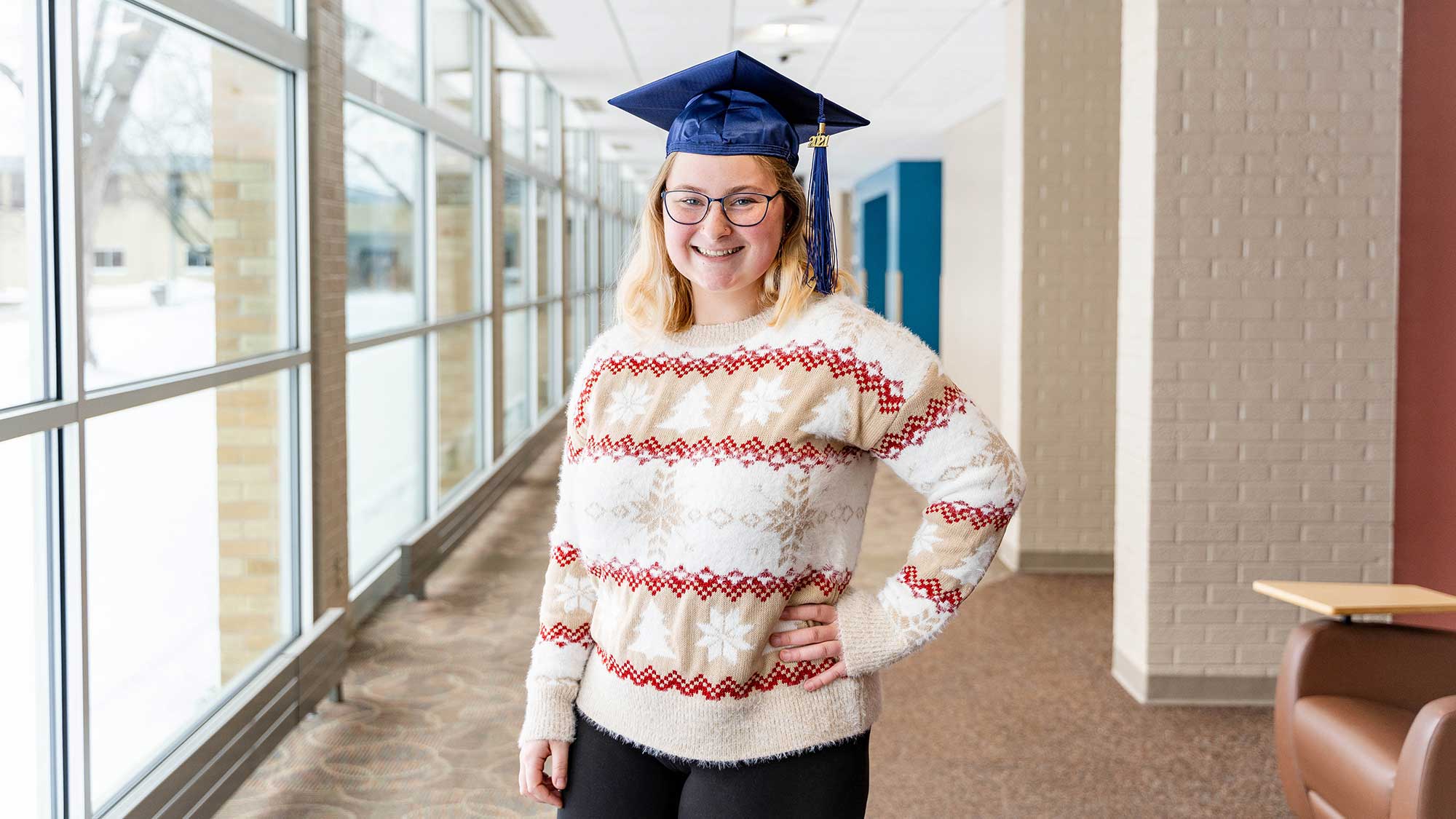 Molly Rydeski standing in an NTC hallway, wearing a festive holiday sweater and a graduation cap.
