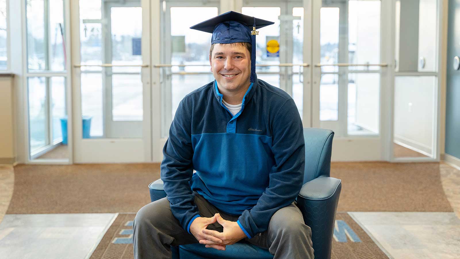 Zachary sitting in a chair while wearing a graduation cap, near the main entrance of the NTC Wausau campus.