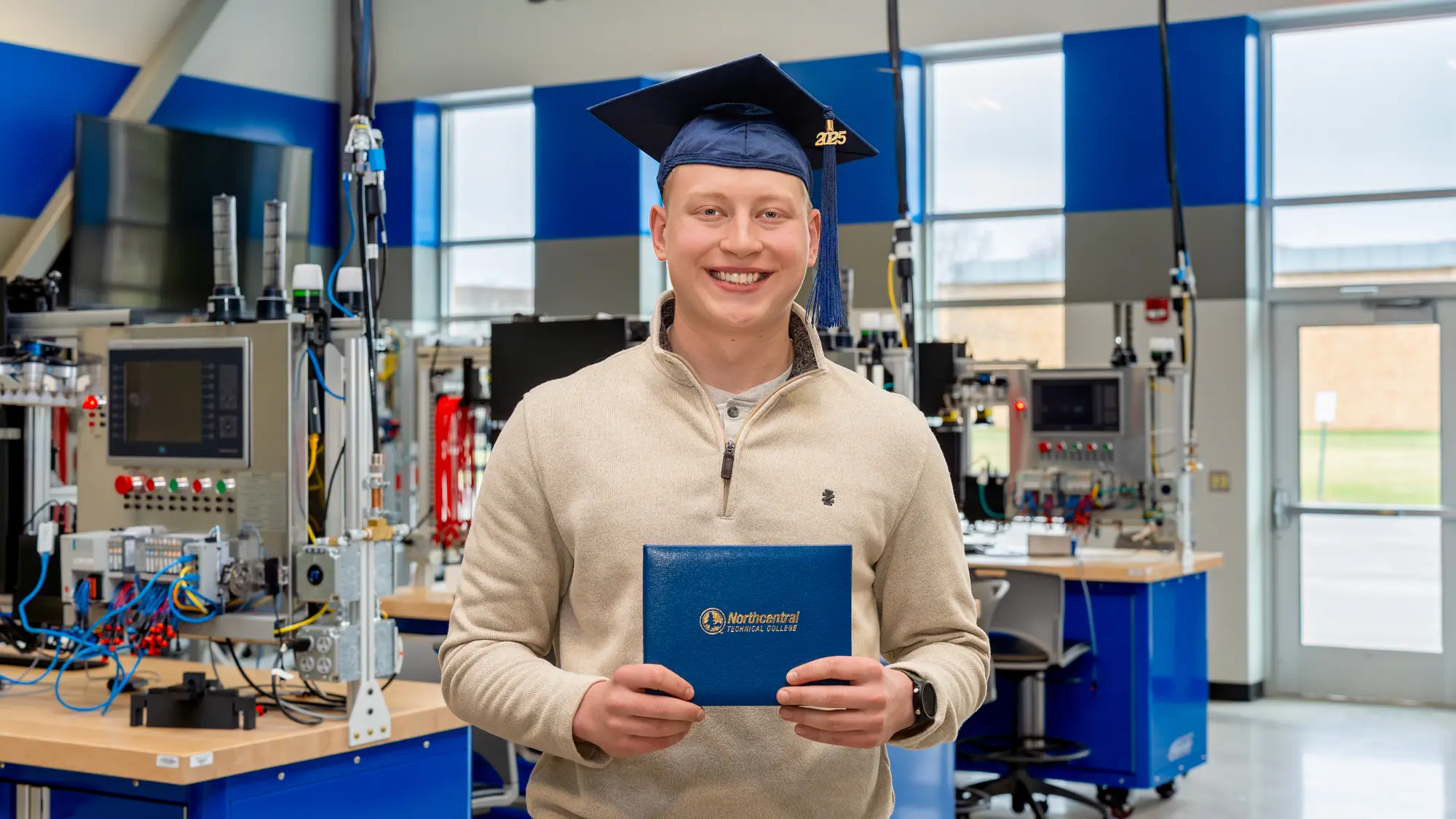 Ethan Sondelski smiling while wearing his graduation cap and holding his diploma, standing inside the Industry 4.0 Smart Manufacturing Lab