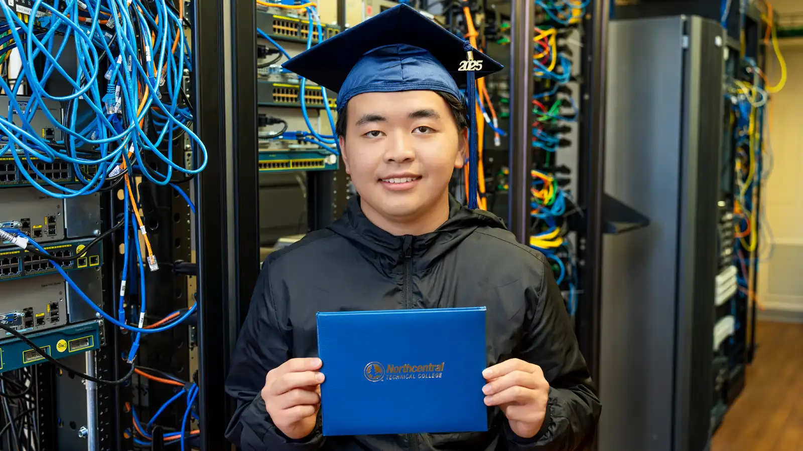 Kong smiling while wearing his graduation cap and holding his diploma, standing in front of some server racks in a network closet.