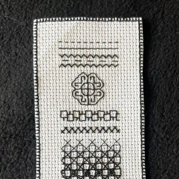 White cross-stitch fabric sample with six rows of black embroidered patterns, including lines, zigzags, a Celtic knot, and geometric shapes, on a dark background.