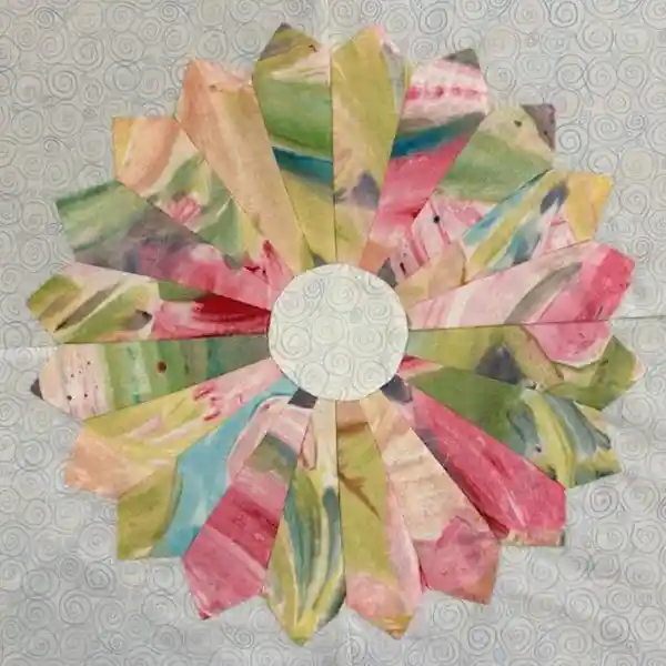 Quilted fabric flower block with a white center and colorful, fan-shaped petals in pink, green, yellow, and blue on a light blue background.