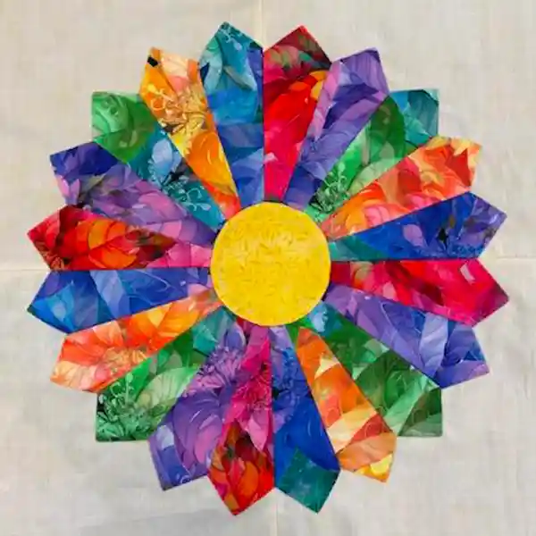 A colorful quilted flower with a bright yellow center and multi-colored petals made from various patterned fabrics on a white background.