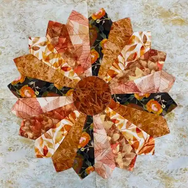Quilted fabric flower with radiating wedge-shaped petals in various autumnal prints, including leaves and florals, on a light background.