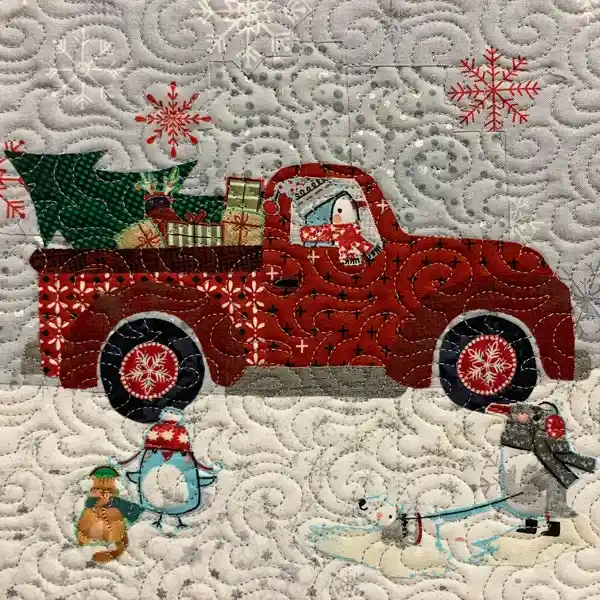 Quilted winter scene: Red truck carries Christmas tree and gifts, driven by a penguin. Penguins, a cat, and person with polar bear are in foreground.