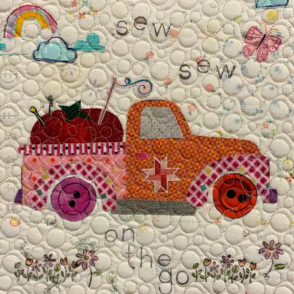 A colorful quilted fabric design shows a pink and orange pickup truck loaded with sewing supplies, surrounded by a rainbow, clouds, and butterflies.