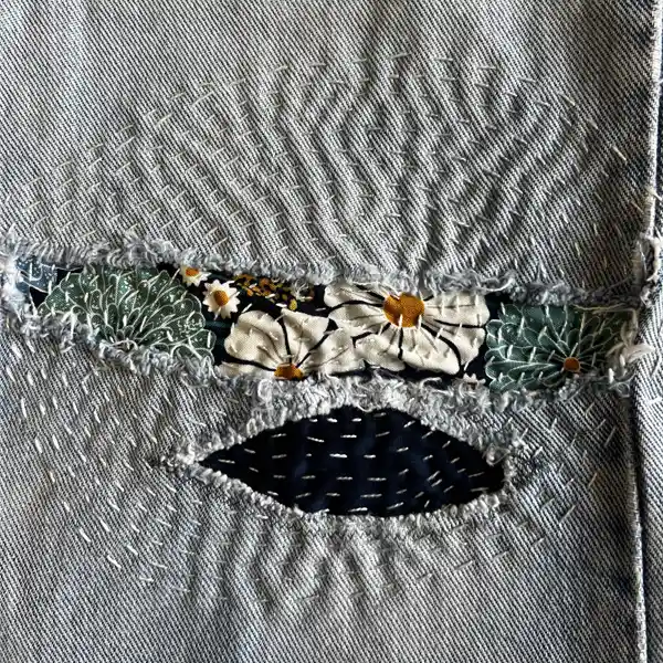 Light blue denim with a horizontal tear revealing white daisy and dark blue patterned fabric, mended with visible white sashiko stitches.