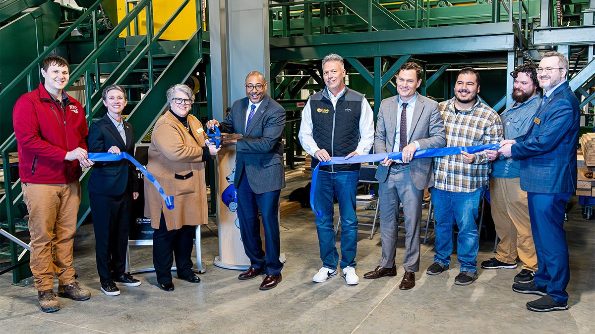 Group of people cutting a blue ribbon at an indoor sawmill facility during a ribbon-cutting ceremony.