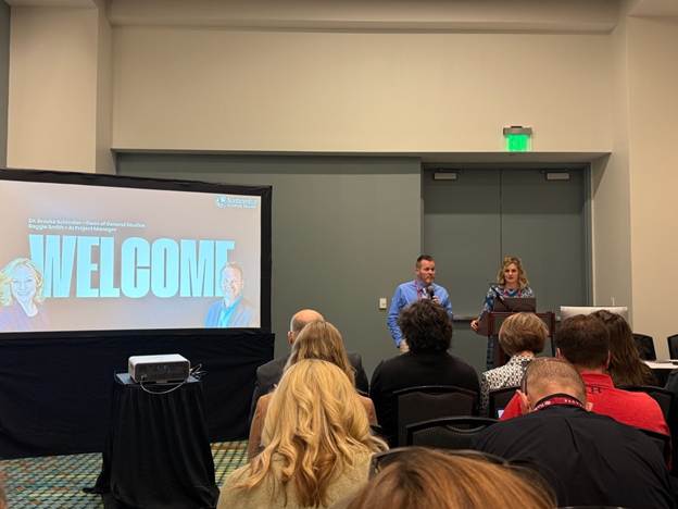 “Two speakers welcome an audience at a conference session with a large screen displaying the word ‘WELCOME.’”