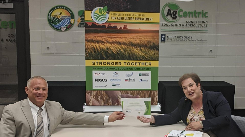 Troy Danielland and Dr. Hara Charlier sit at a table and display a memorandum of understanding (MOU)