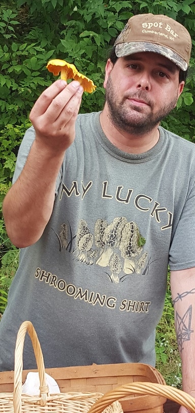 Tavis standing outside with trees behind him, while holding some mushrooms in his right hand while wearing a shirt that reads My Lucky Shrooming Shirt