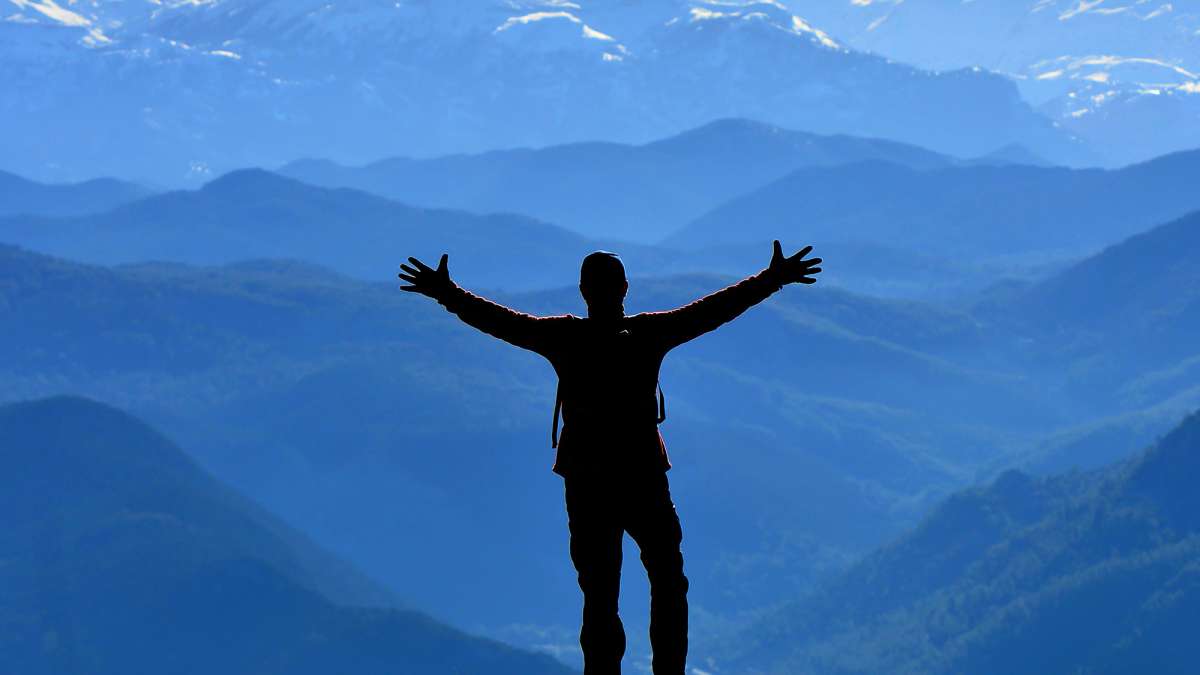 A man stands at the top of a hill looking out at the mountains with his hands spread apart in the air