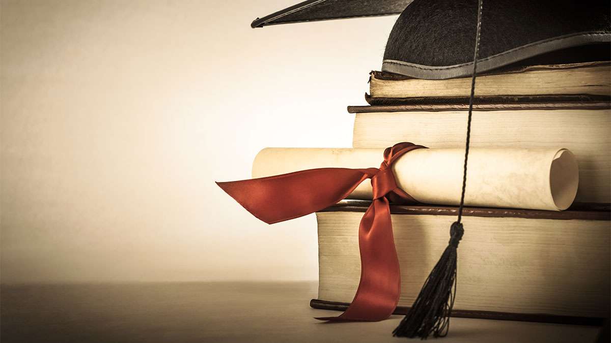 A graduation hat sits on top of three large books and a rolled up paper wrapped in a red bow