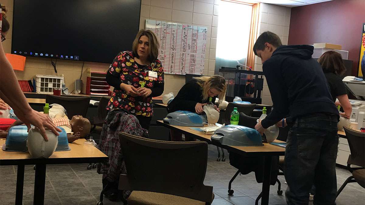 Several students are at tables standing over medical training mannequins. An instructor is in the classroom observing them