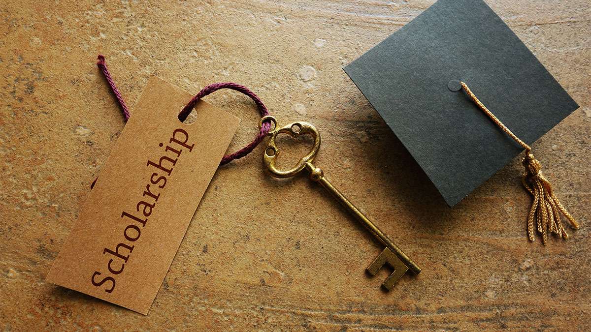 A golden key with a tag that says scholarship is placed next to a graduation hat on a wood table