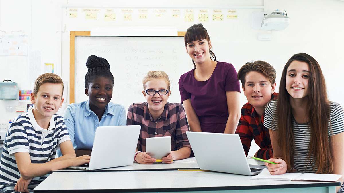 Several smiling students of varying ages sit together at a table with two laptops and a tablet inside of a classroom