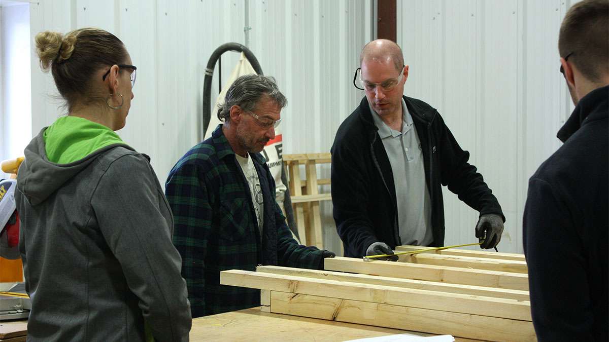 Several people stand next to a table inspecting wooden boards. One man is taking measurements of the boards with a measuring tape..