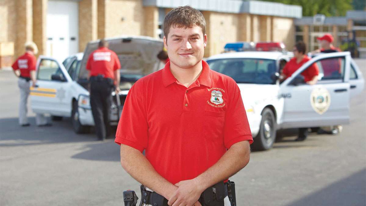 A Sheriff's Academy student stands with his hands on his belt in front of two squad cars