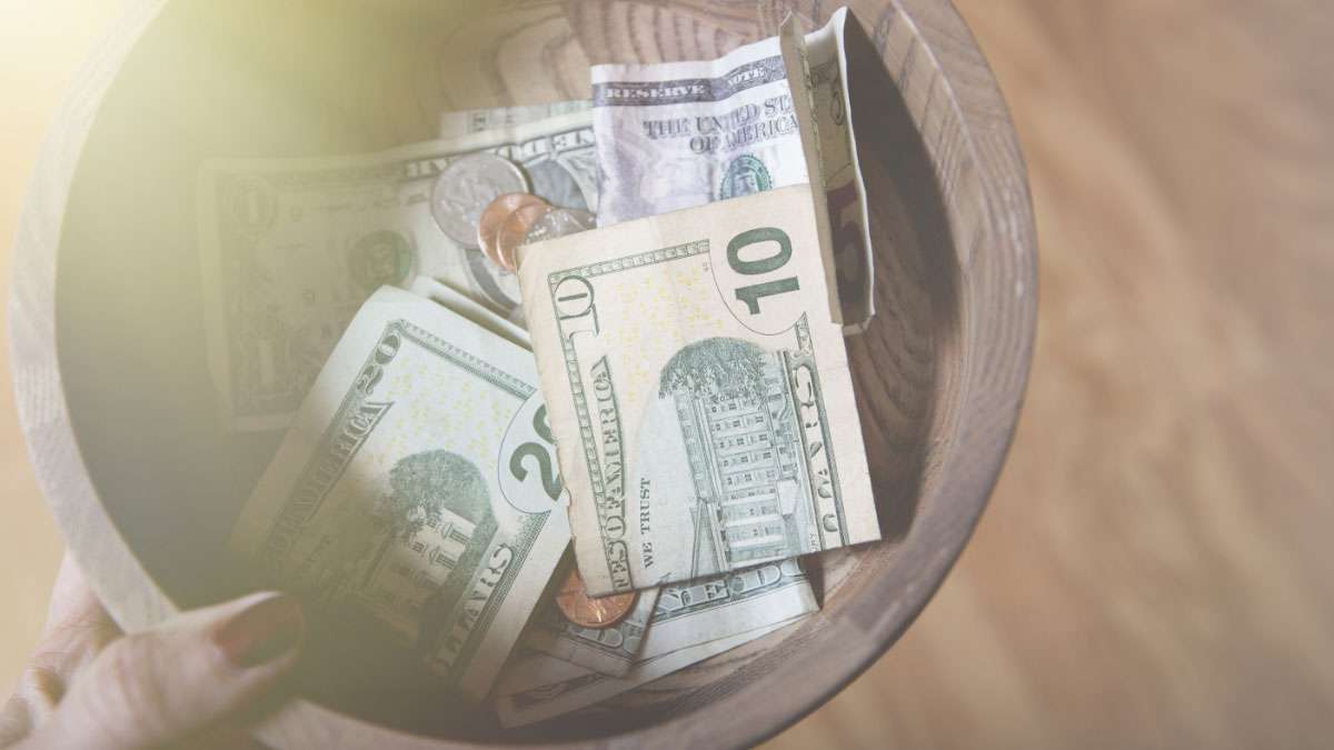 A person is holding a collection bowl with coins and dollar bills in it