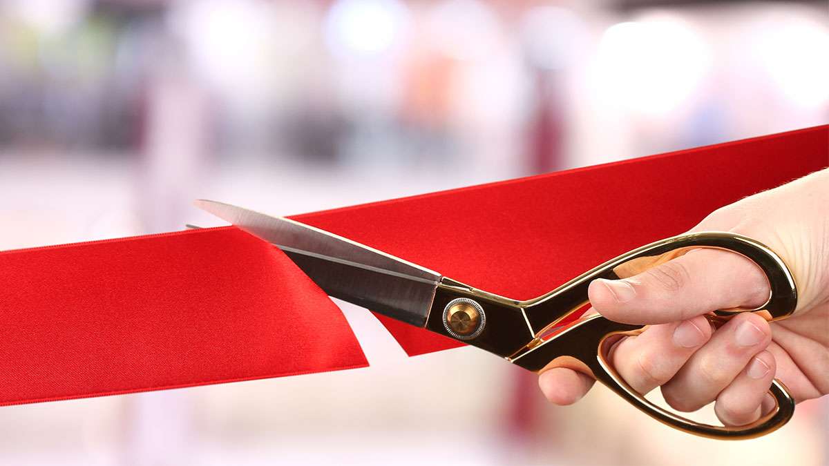 A hand is holding a pair of scissors cutting a large red ribbon