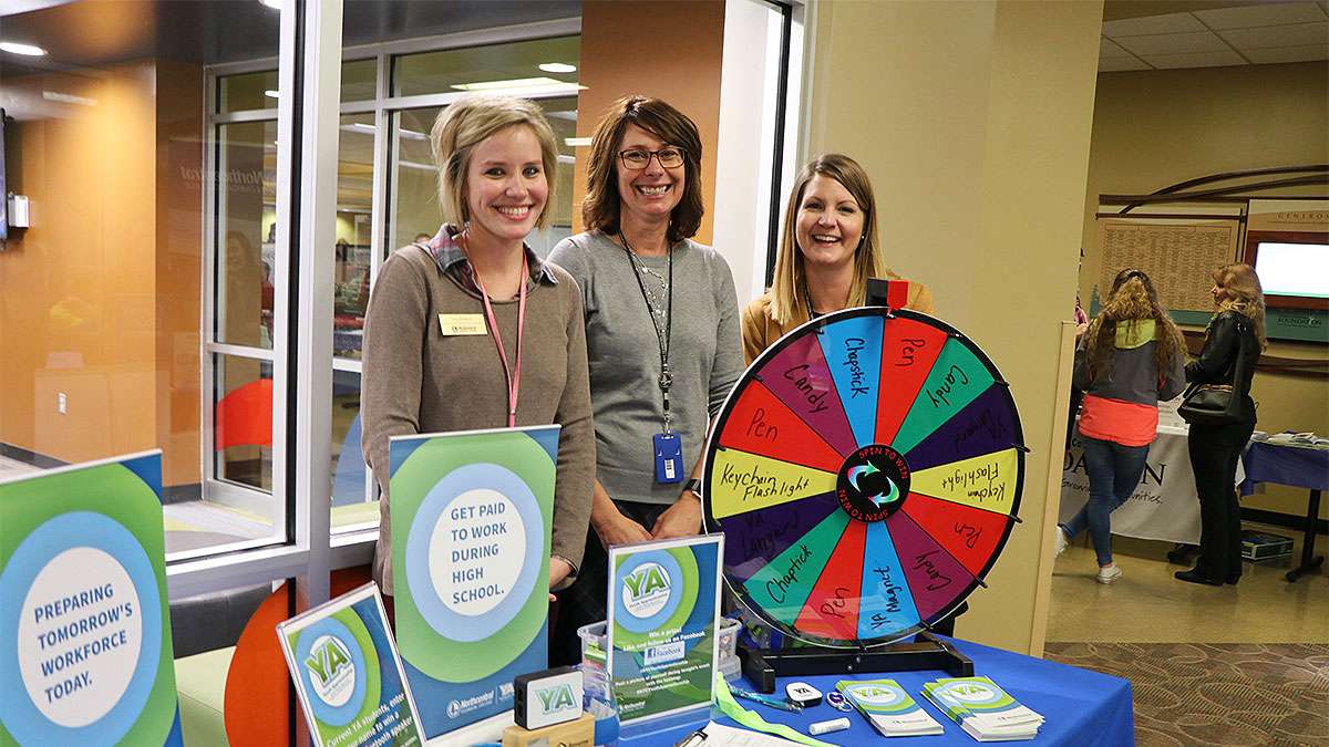 3 people stand behind promotional material for NTC's Open House