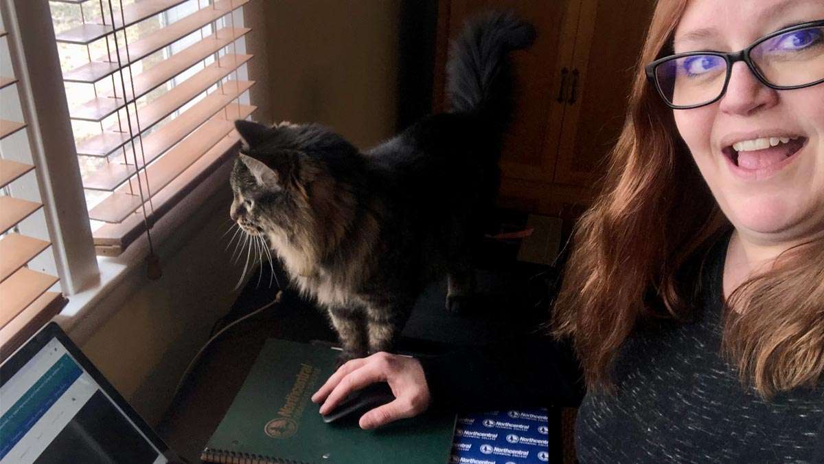 Laura working from home with her cat sitting on her desk next to her.