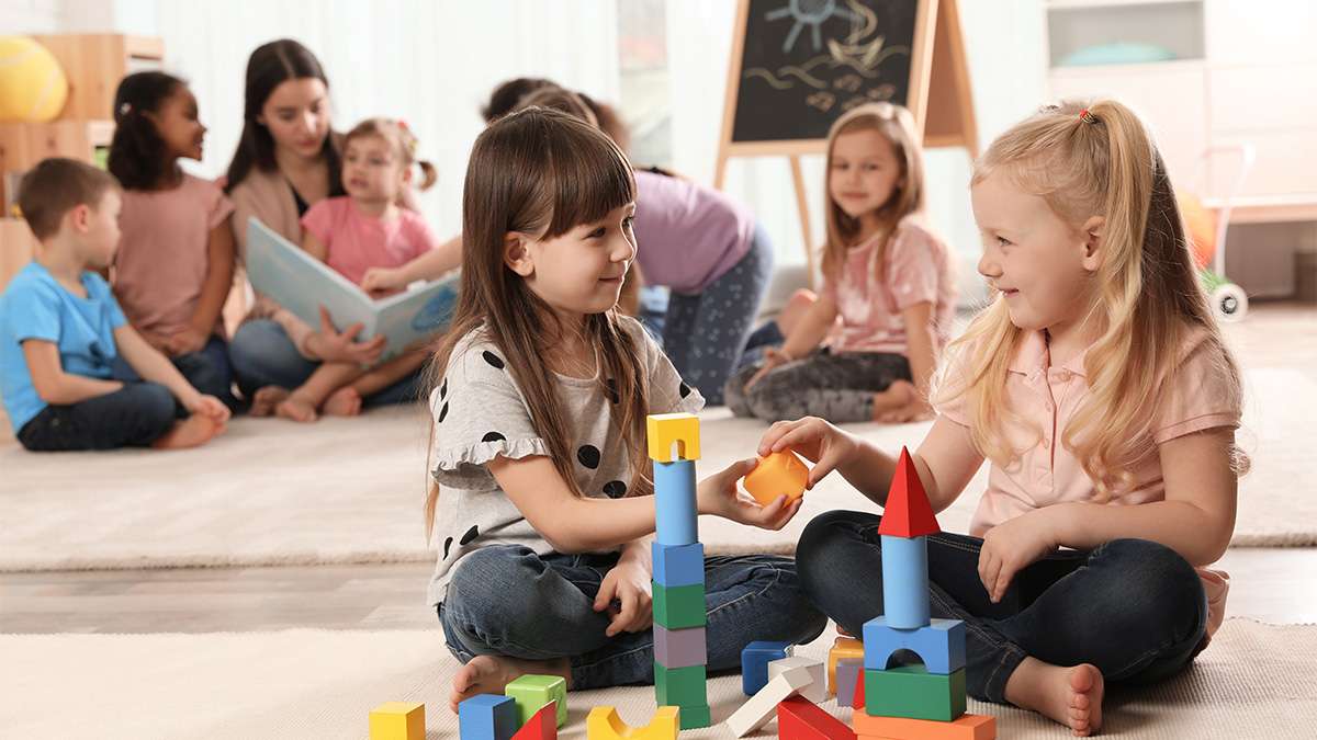 Children are playing with toy blocks and having a story read to them by a child care worker
