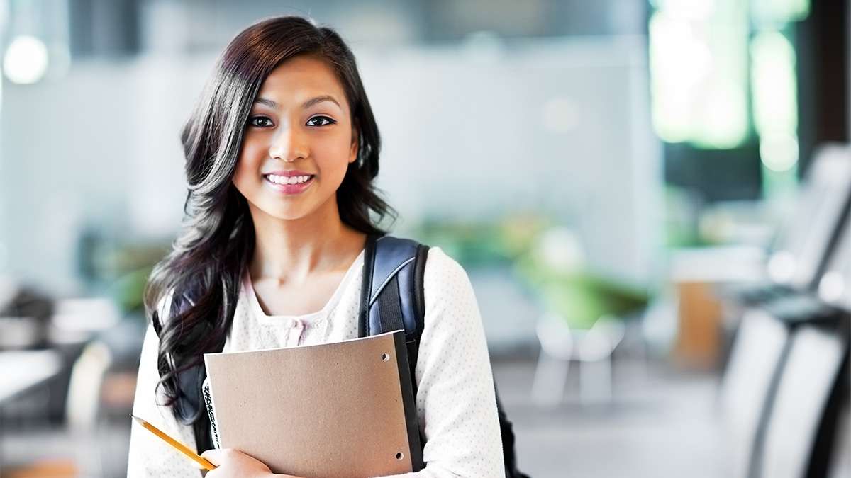 A student stands in a classroom with a pencil in hand and a notepad in her arms.