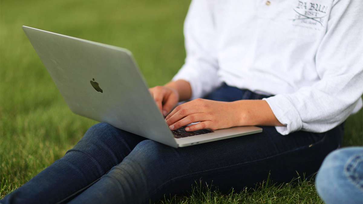 A student is sitting on the grass typing on an Apple Macbook Pro laptop.