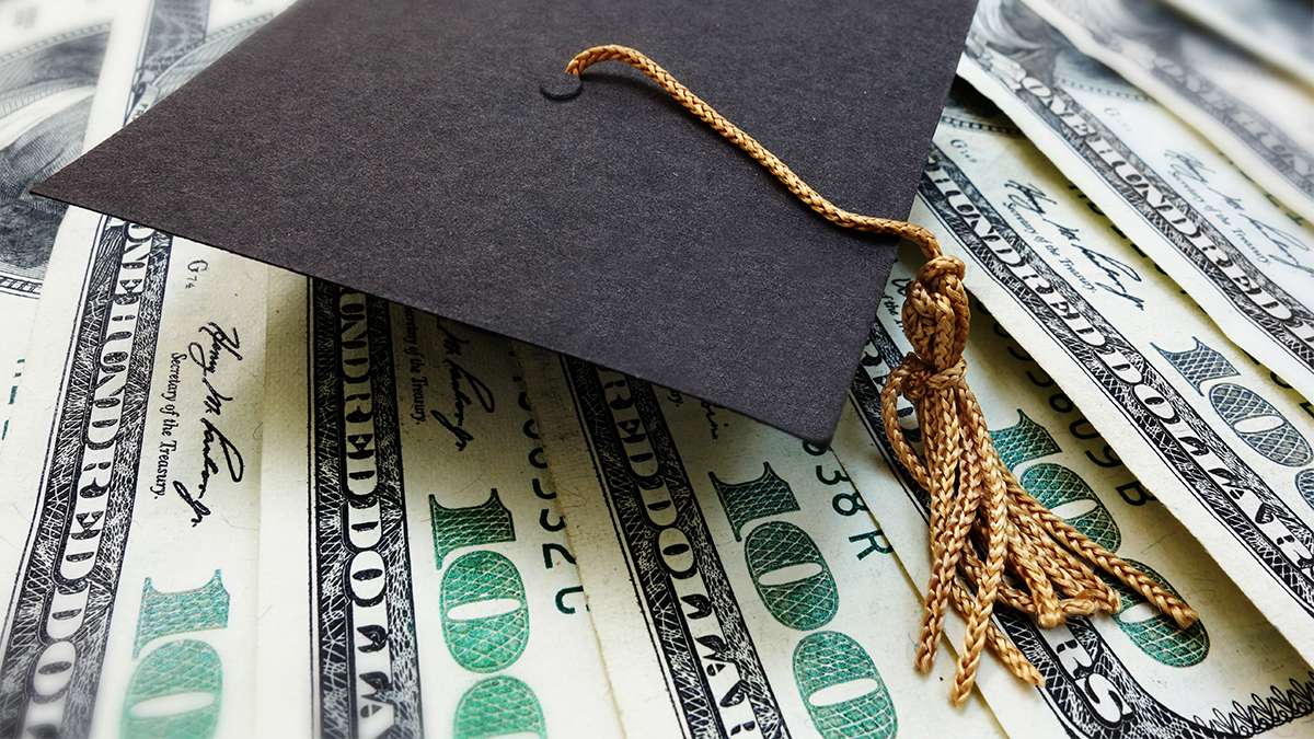 A black graduation hat with gold tassels sits on top of several one-hundred dollar bills.