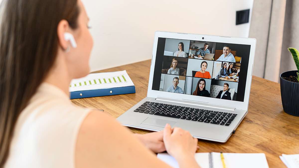 A woman using earbuds and a laptop at a desk, is attending an online meeting with several other people.