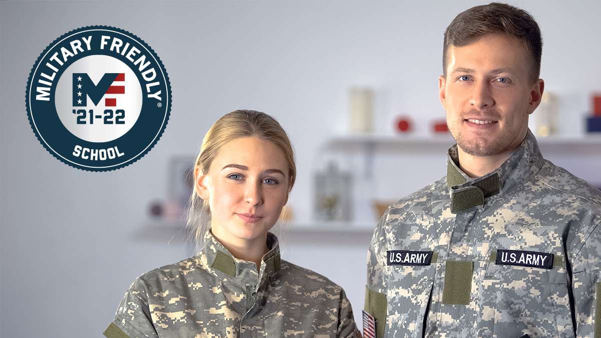 Two military soldiers stand side by side in uniform with the American flag behind them.