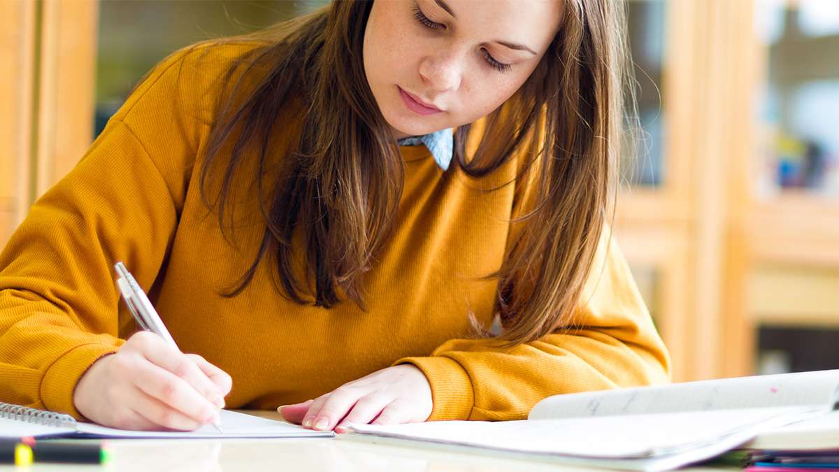 A young college student in sits at a table with open books writing notes down. 