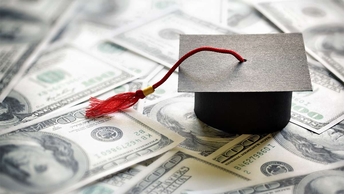 A black college graduation cap with red tassels sits on top of one-hundred-dollar bills.