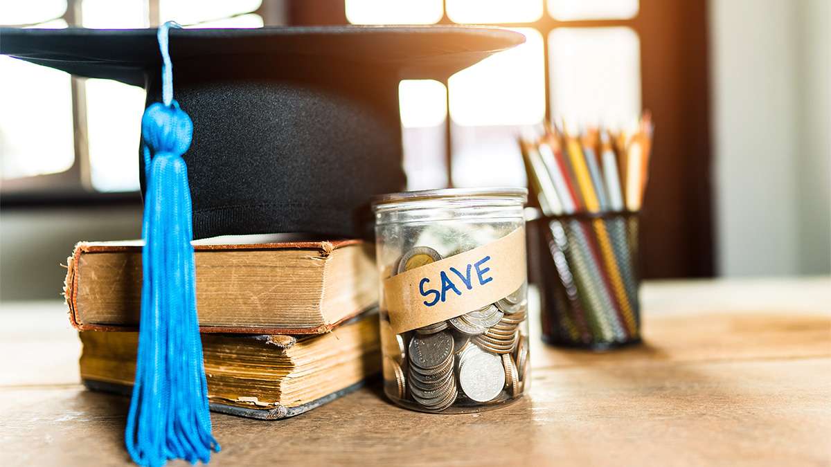 A graduation hat is placed next to a jar full of pencils, a couple of textbooks, and a jar full of coins.