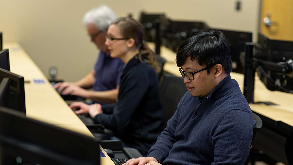 Three students in a classroom are typing on keyboards at desktop workstations.