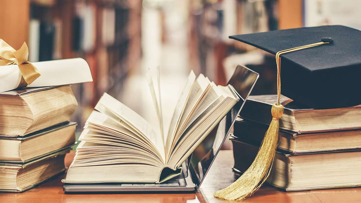 Several books, a laptop, and a graduation hat are stacked on top of a table inside of a library.