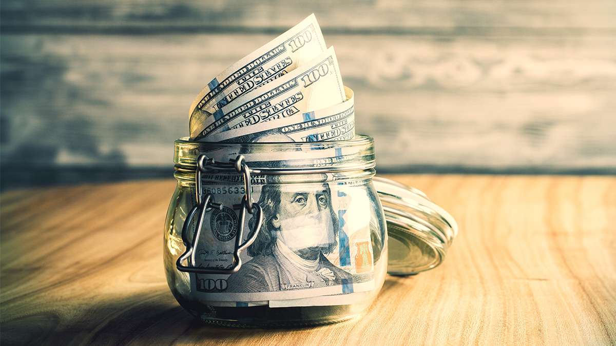 An open jar on top of a wooden table filled with one-hundred dollar bills.