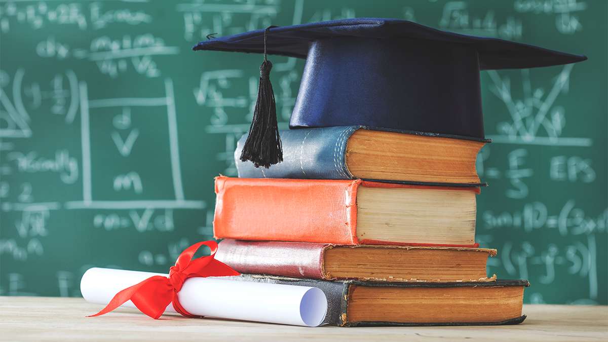 A stack of books underneath a graduate hat in front of green chalkboard with math written on it.