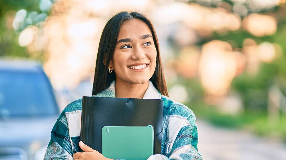 A young woman holding a binder and notepad in her arms looks up towards the sky.