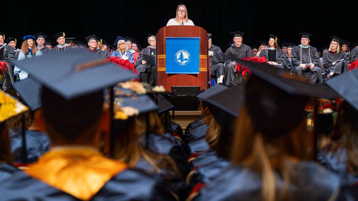 Crowd of NTC graduates seated wearing black mortarboards with gold accents, viewed from behind toward a podium and stage.
